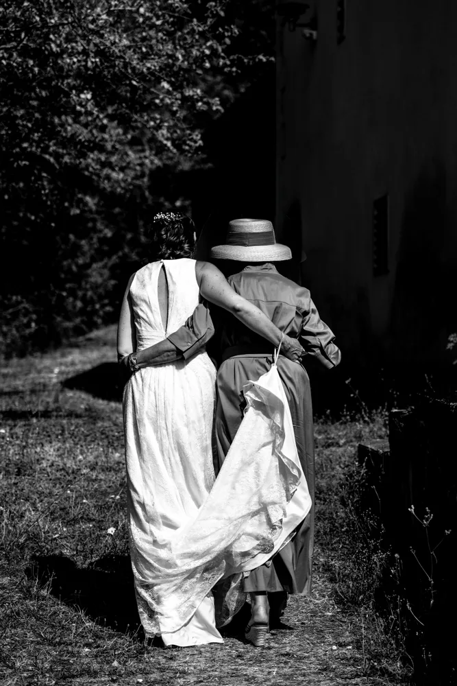 Une mariée s’éloigne, enlacée à une proche qui tient sa traîne, en noir et blanc.